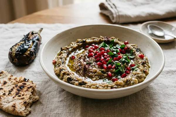 Schale rauchige Baba-Ghanoush-Auberginencreme mit Olivenöl, Granatapfelkernen und Petersilie, daneben eine verkohlte Aubergine und Fladenbrot