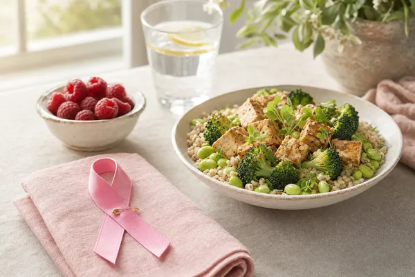 Rosa Schleife als Zeichen der Brustkrebs-Aufklärung auf einer rosa Leinenserviette, dahinter eine Schale mit gebratenem Tofu, Brokkoli, Edamame und Graupen, ein Schälchen Himbeeren und ein Glas Wasser mit Zitrone