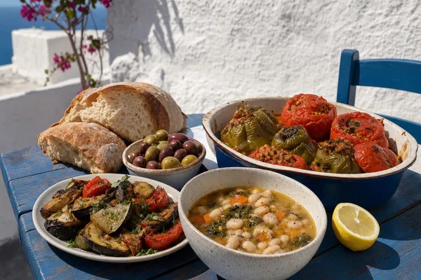 Sommerlicher Taverna-Tisch auf einer griechischen Insel: Schalen mit Fasolada, gefüllten Tomaten, geschmortem Ofengemüse und Oliven auf blau gestrichenem Holztisch vor weiß getünchter Mauer im Mittagslicht